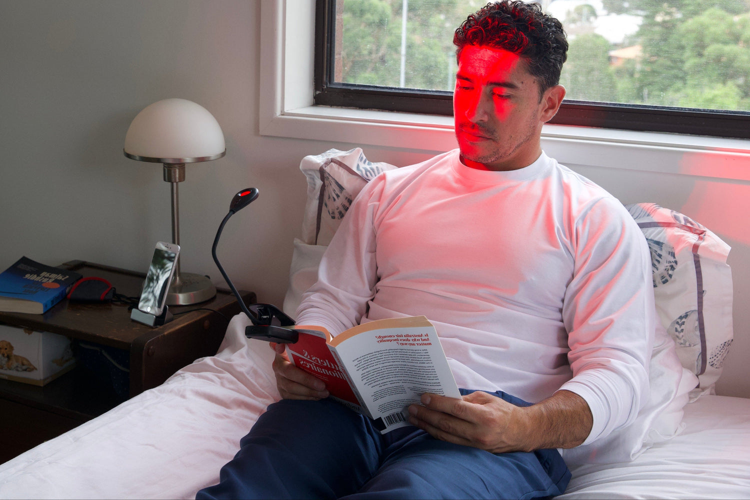 Man sitting on a bed reading a book with a red light on his face, in a room with a window and lamp.