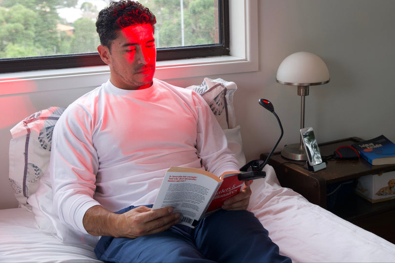 Man sitting on a bed reading a book with a red light on his face, in a room with a window and lamp.