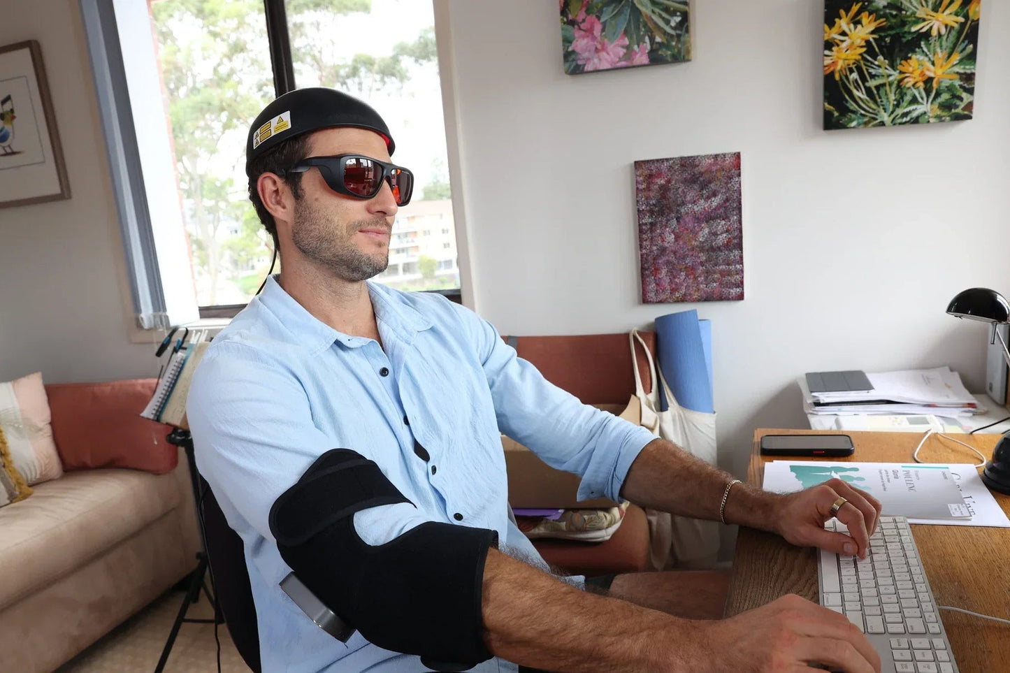 Man wearing the HeroHat Laser cap, protective glasses and an elbow wrap at a laptop
