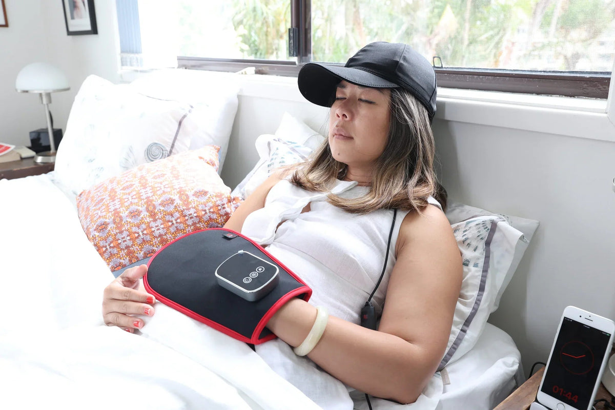 Woman resting in bed while using the HeroHat LED cap and a red light therapy belt