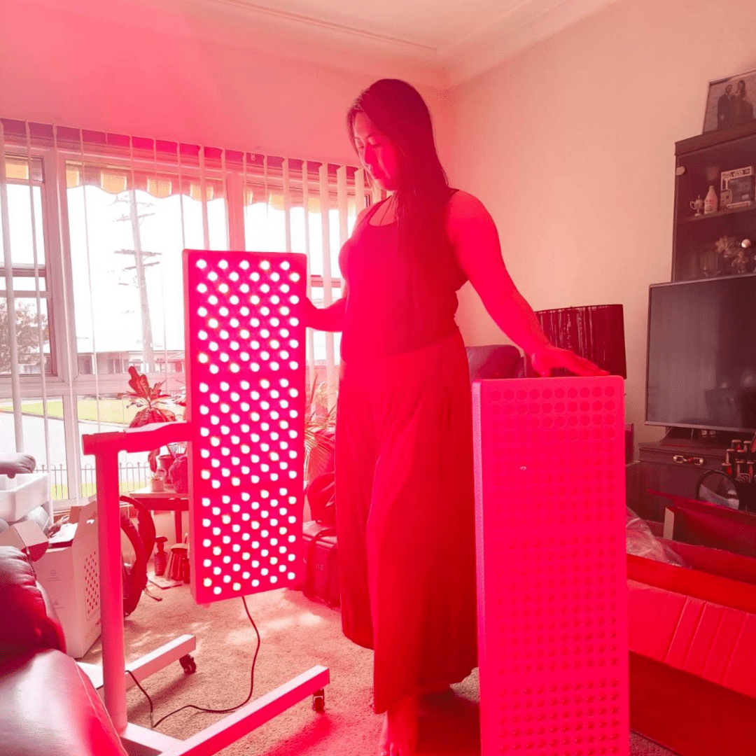 Woman standing beside an illuminated Hero300 red light therapy panel by a window